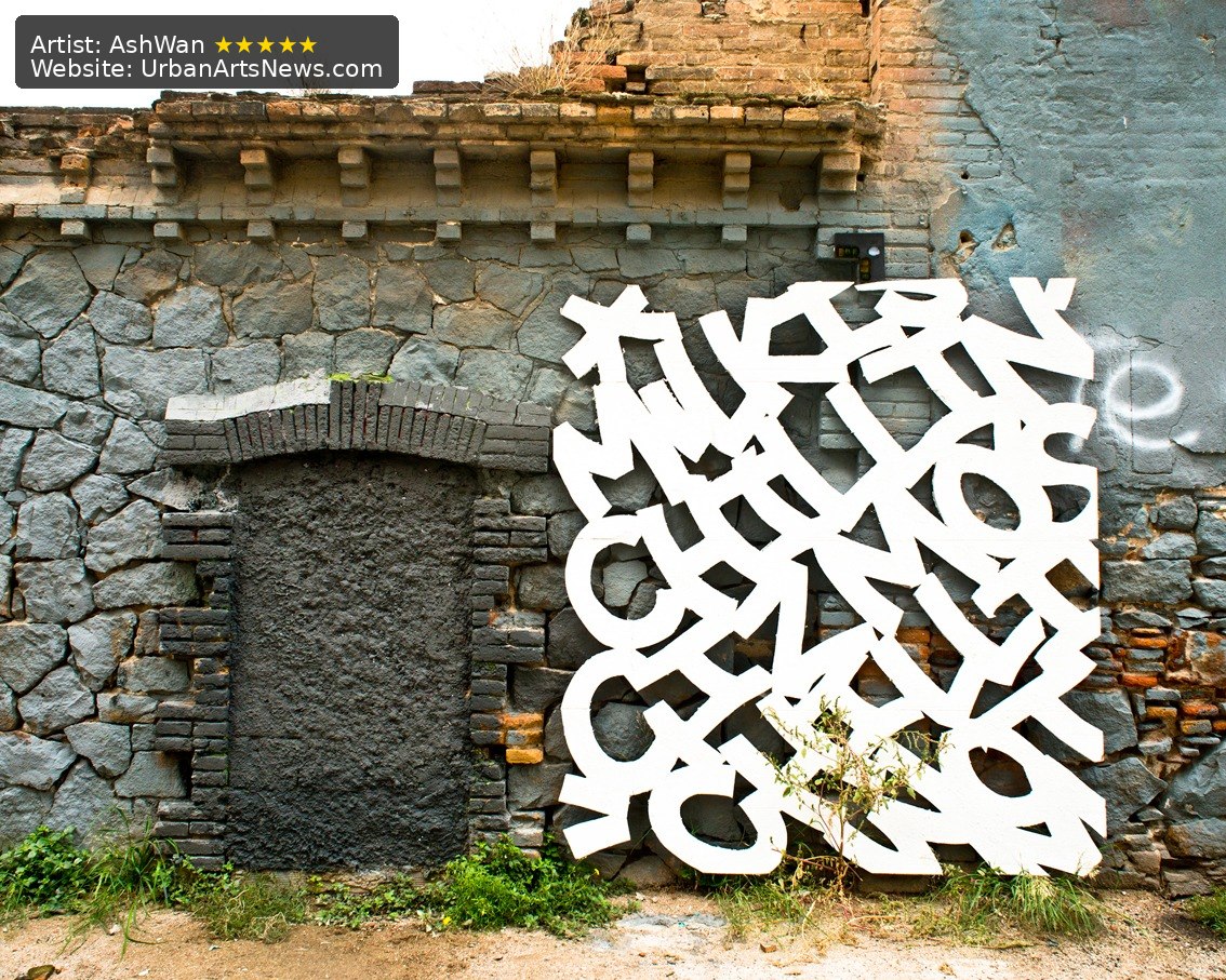 A freestanding white abstract letter installation by AshWan placed against a weathered urban brick wall.
